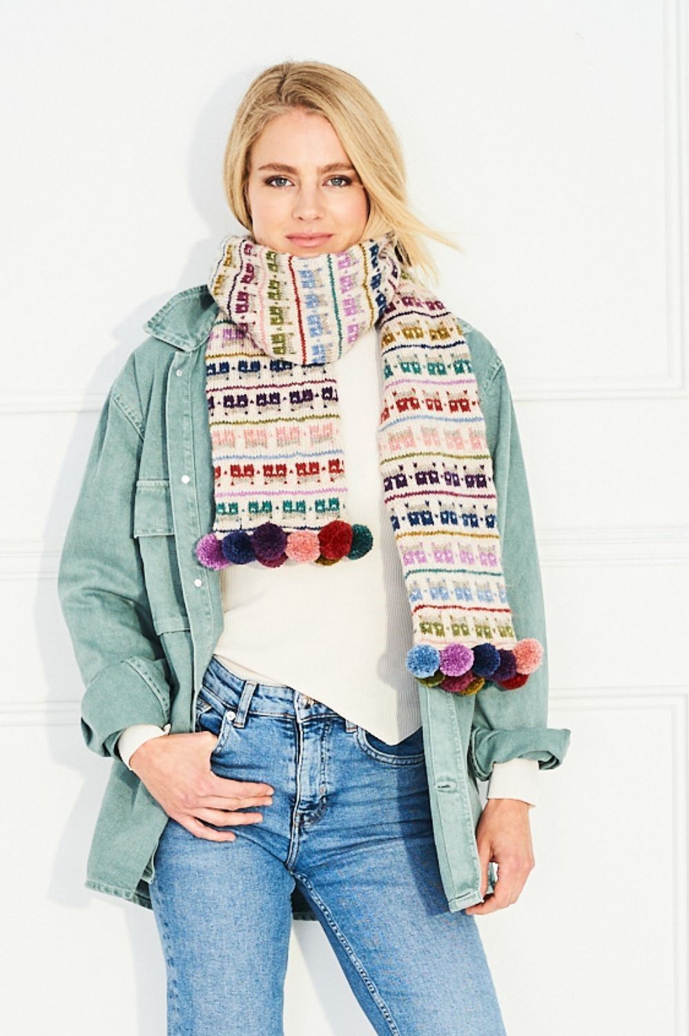 A woman is wearing a cream knitted scarf with pink, purple, red, green and blue colourwork houses.