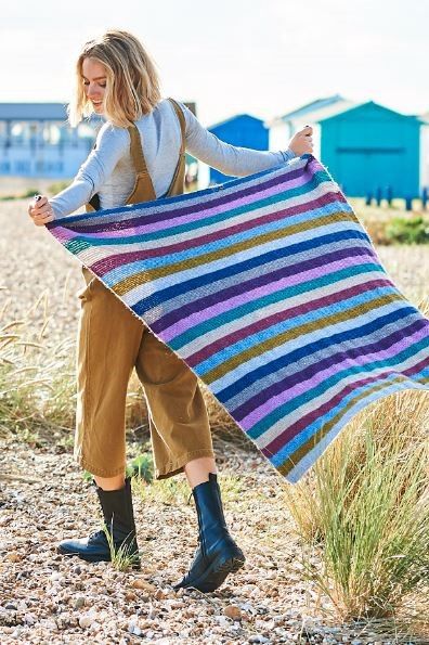 A woman is holding a stripy knitted blanket in blues, greens, purples, pinks and greys.