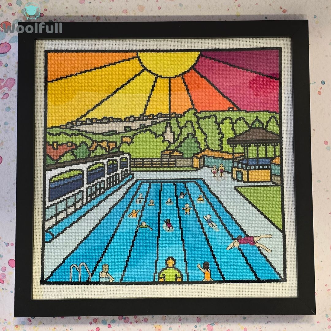 A cross stitch in a black frame. The cross stitch is a cartoon style picture of Hathersage Pool in the Peak District.