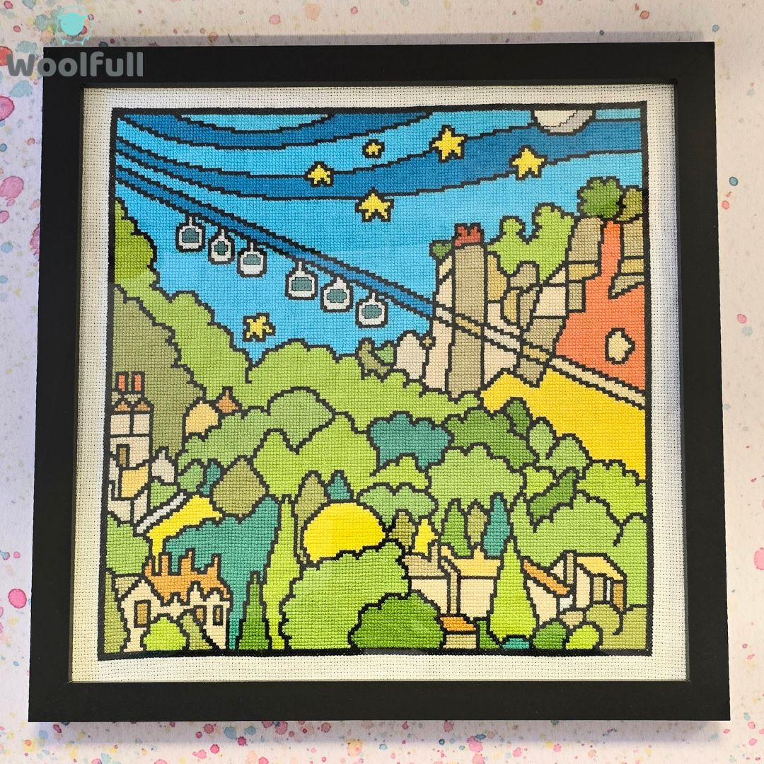 A cross stitch in a black frame. The cross stitch is a cartoon style picture of the Heights of Abraham in the Peak District.