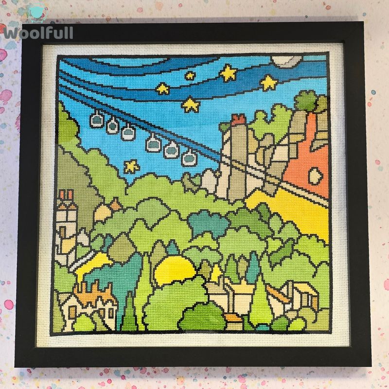 A cross stitch in a black frame. The cross stitch is a cartoon style picture of the Heights of Abraham in the Peak District.