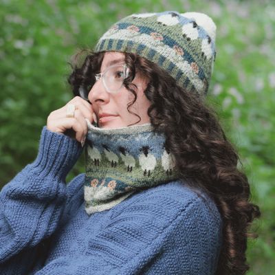 A woman is wearing a blue and green knitted hat and matching cowl, with colourwork sheep.
