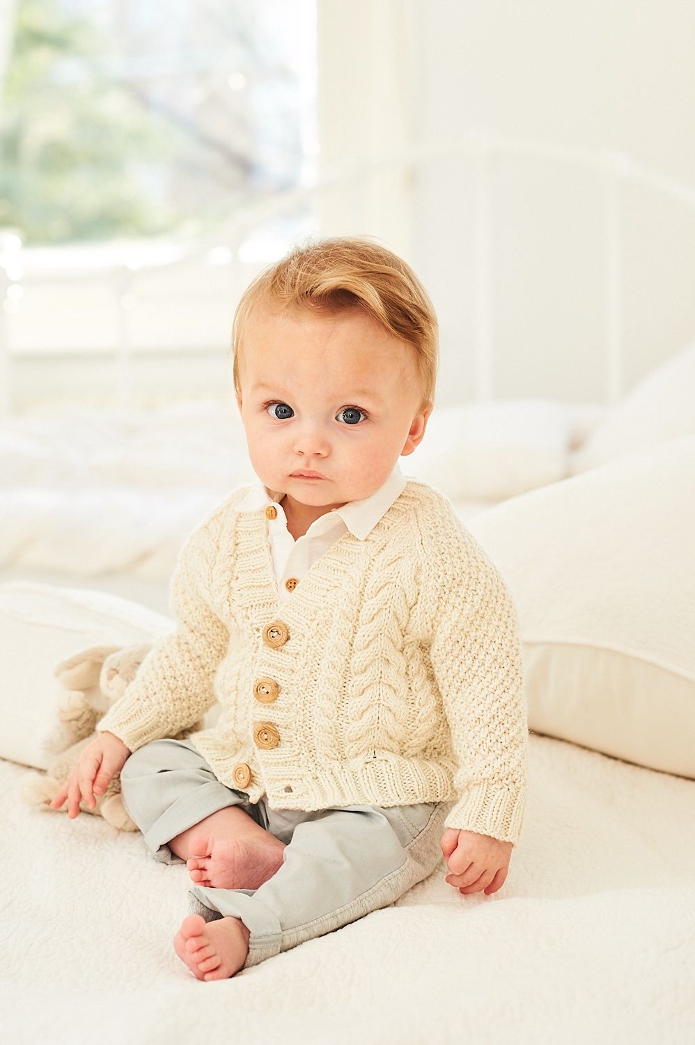 A baby is wearing a cream cable knit cardigan.