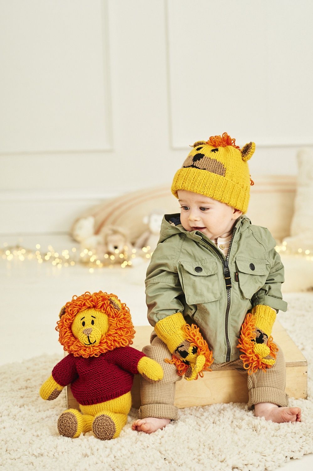 A baby is wearing a yellow knitted lion hat and matching mittens, while sitting next to a knitted toy lion in a red sweater.