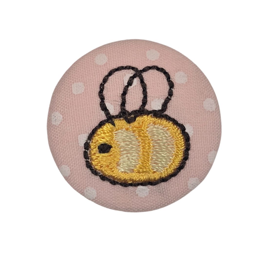 A round, pink fabric covered button with an embroidered bee.