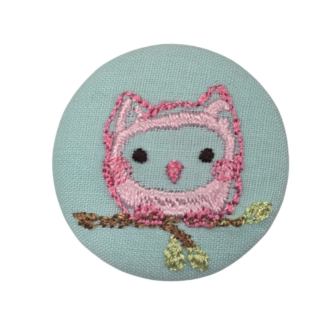 A round, blue fabric covered button with a pink embroidered owl.