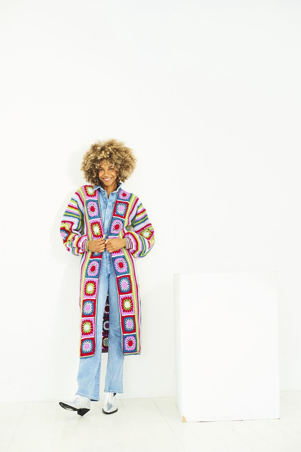 A woman is wearing a crocheted coat in shades of red, pink, blue, white and green.