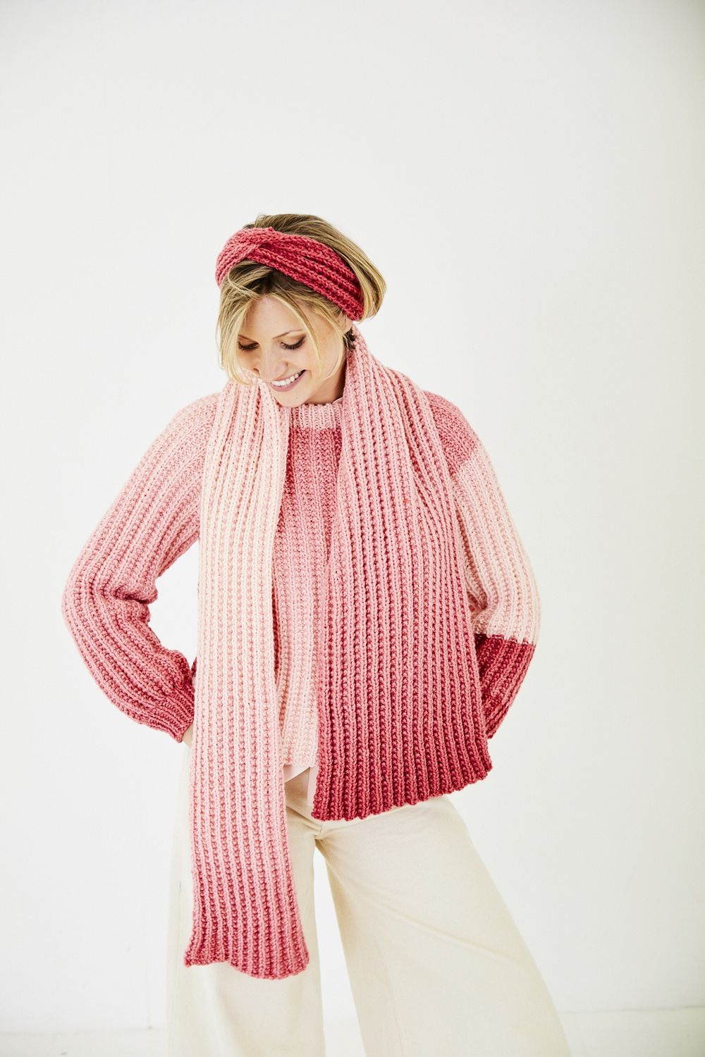 A woman is wearing a knitted sweater in a pink gradient yarn with matching headband and scarf.