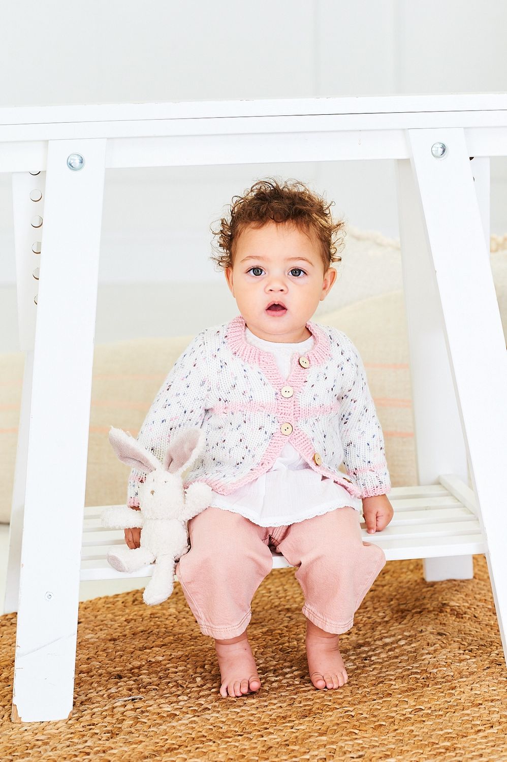 A baby is wearing a knitted cardigan in white with colourful speckles and pink edging.