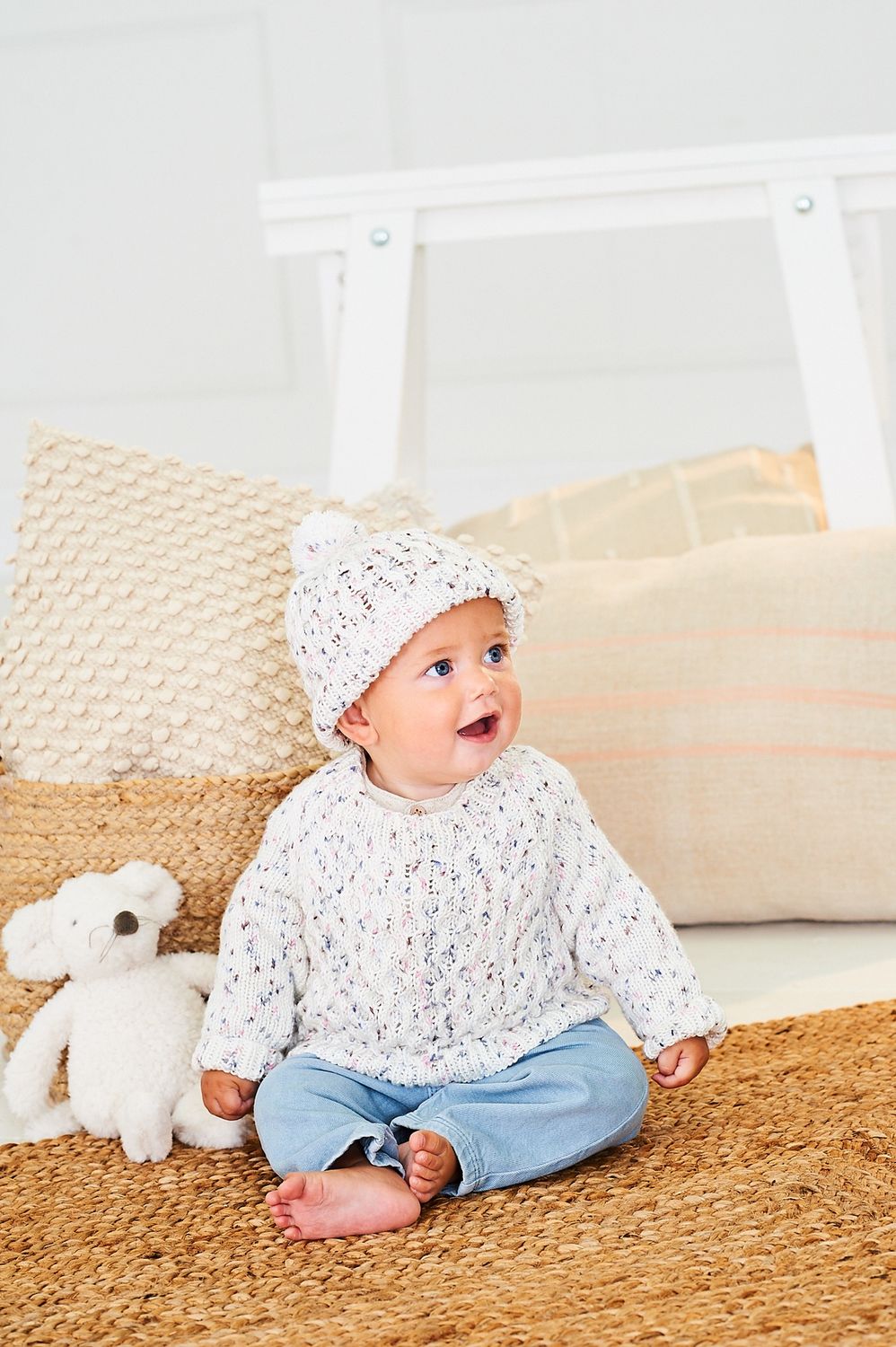 A baby is wearing a knitted sweater and hat in a white yarn with colourful speckles.