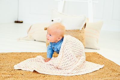 A baby is wearing a knitted blanket in a cream yarn with colourful speckles.