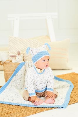 A baby is wearing a knitted sweater and hat in a white yarn with colourful flecks and blue trim, along with matching blanket.
