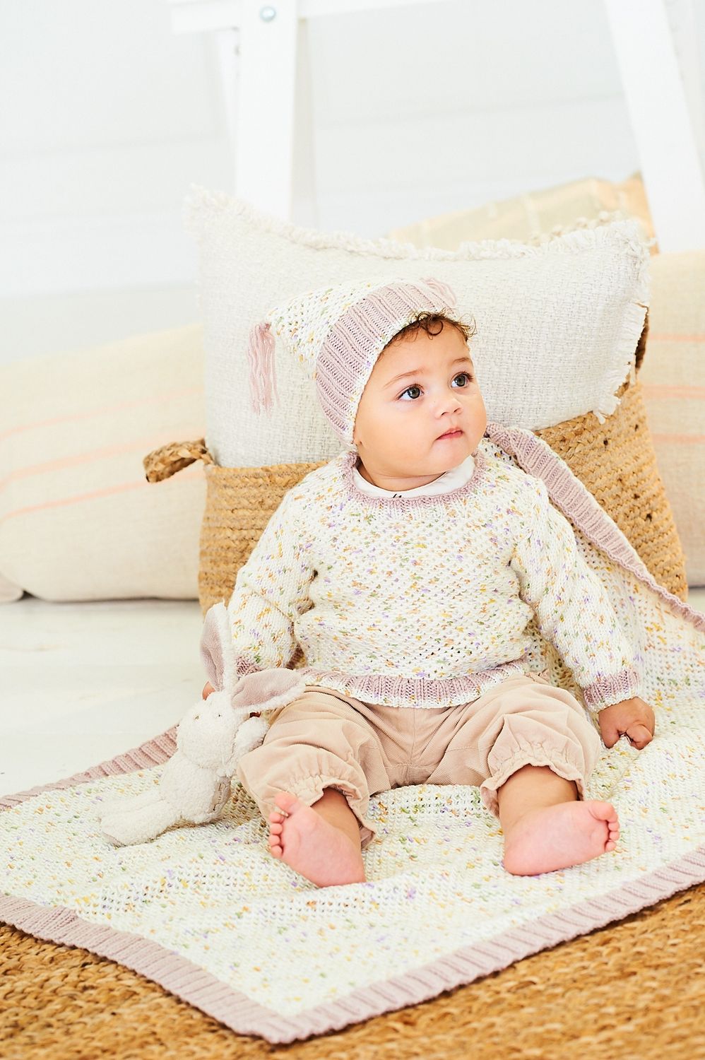 A baby is wearing a knitted sweater and hat in a cream yarn with colourful flecks and pink trim, along with matching blanket.