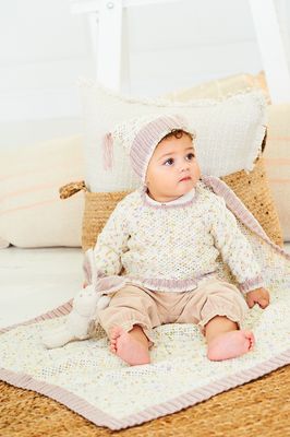 A baby is wearing a knitted sweater and hat in a cream yarn with colourful flecks and pink trim, along with matching blanket.