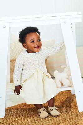 A baby is wearing a cream crocheted cardigan.