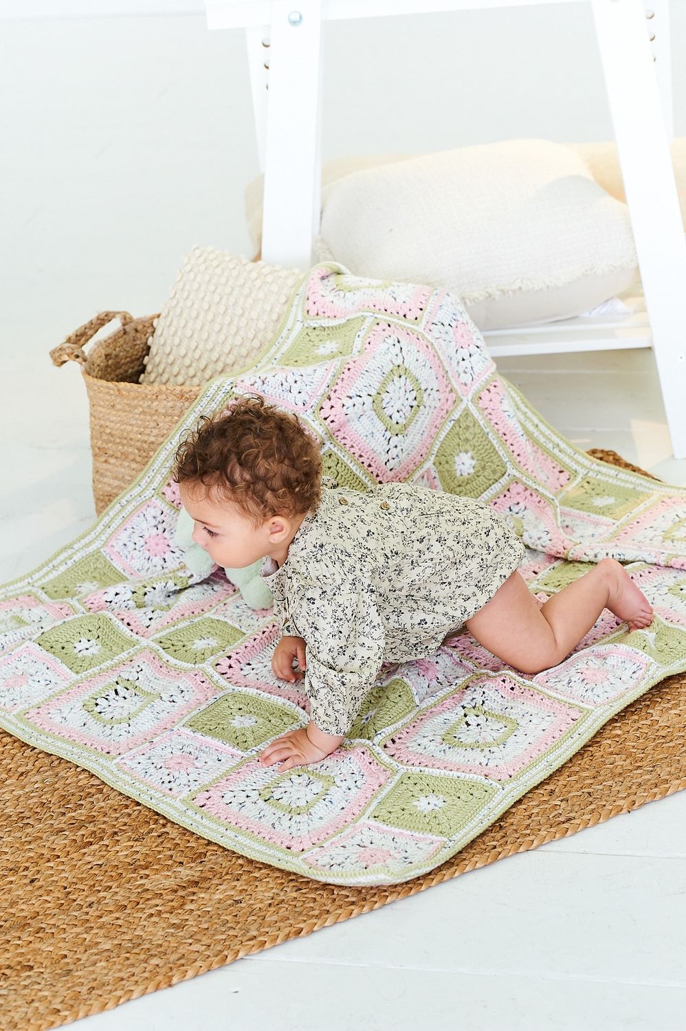 A baby is crawling on a crocheted blanket, made up of green, pink and white squares.