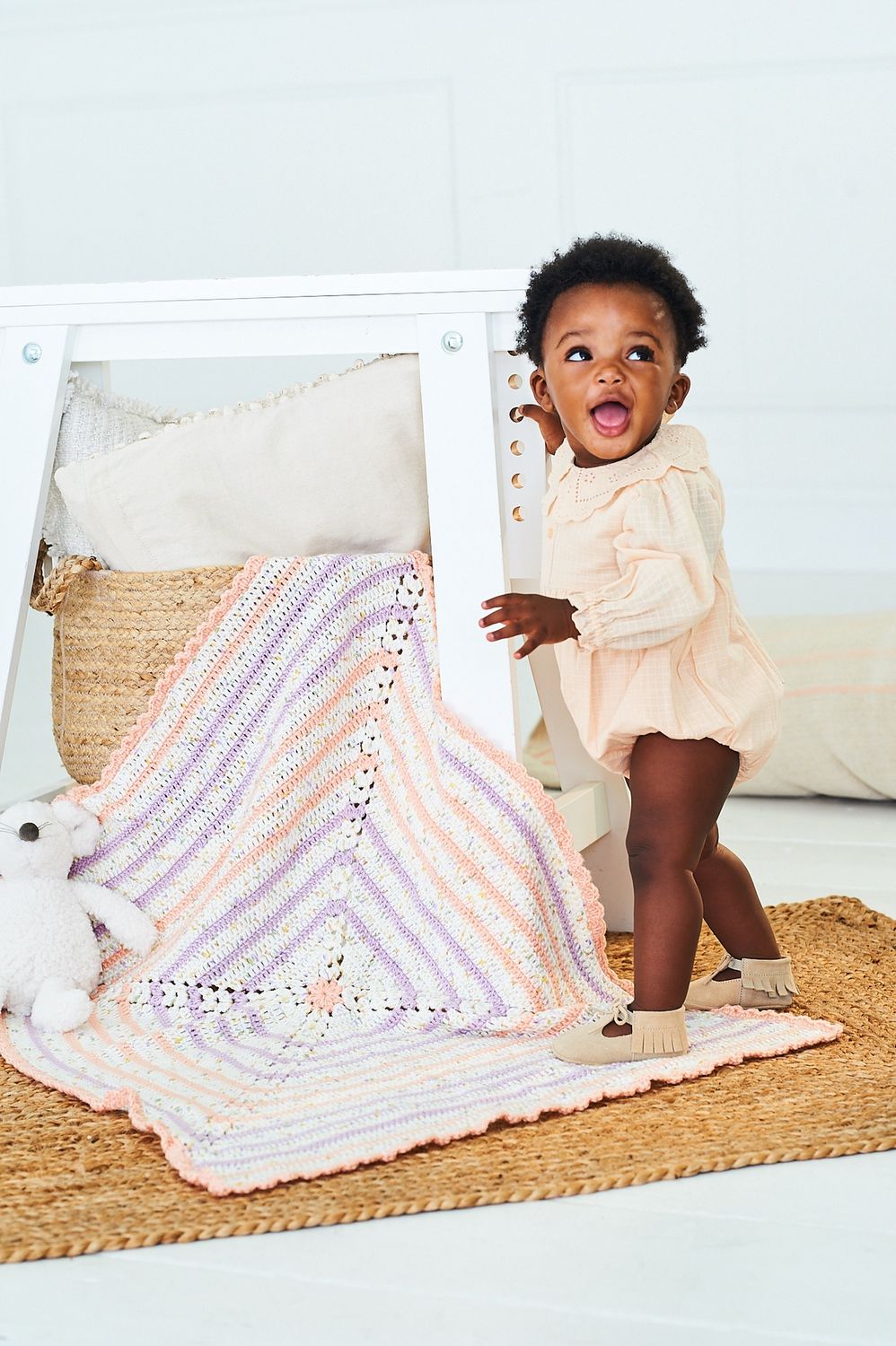 A baby is standing on a crocheted blanket, with peach, purple and white stripes.