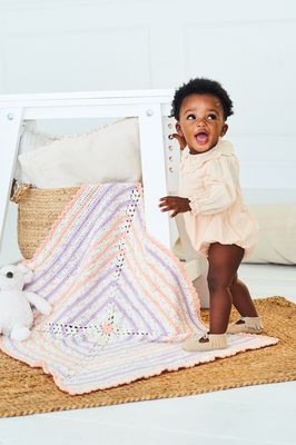 A baby is standing on a crocheted blanket, with peach, purple and white stripes.