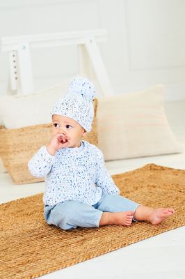 A baby is wearing a crocheted sweater and matching hat in a white yarn with small blue flecks.