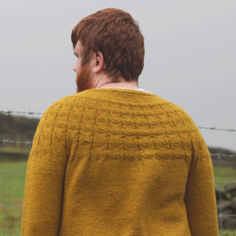 A man is wearing a yellow knitted sweater with textured stitches on the yoke.