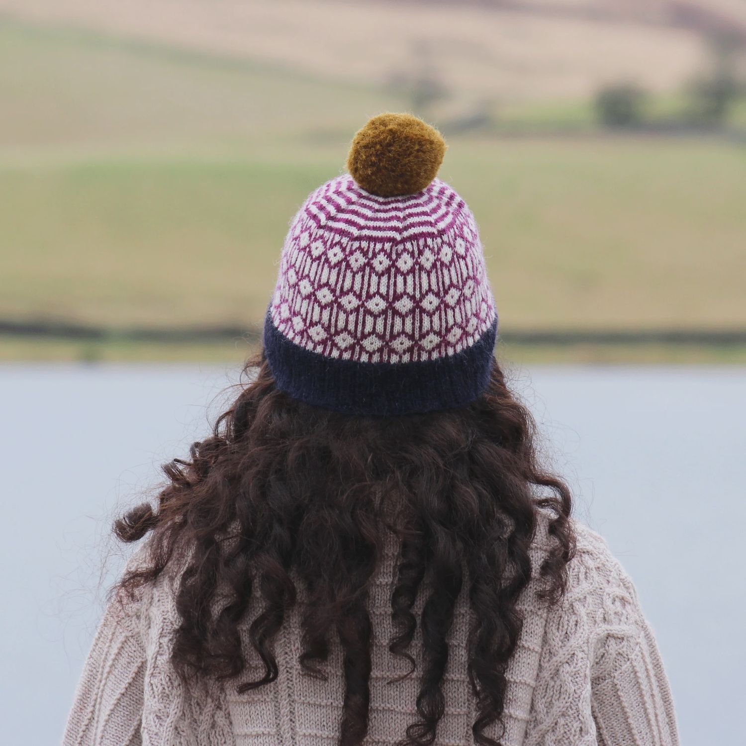 A woman is wearing a knitted colourwork hat in navy blue, purple and cream with a mustard pompom.