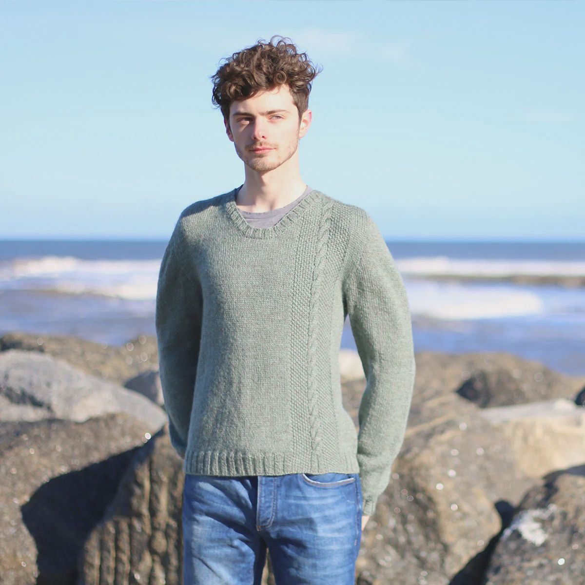 A man is standing in front of rocks, wearing a pale grey knitted sweater.