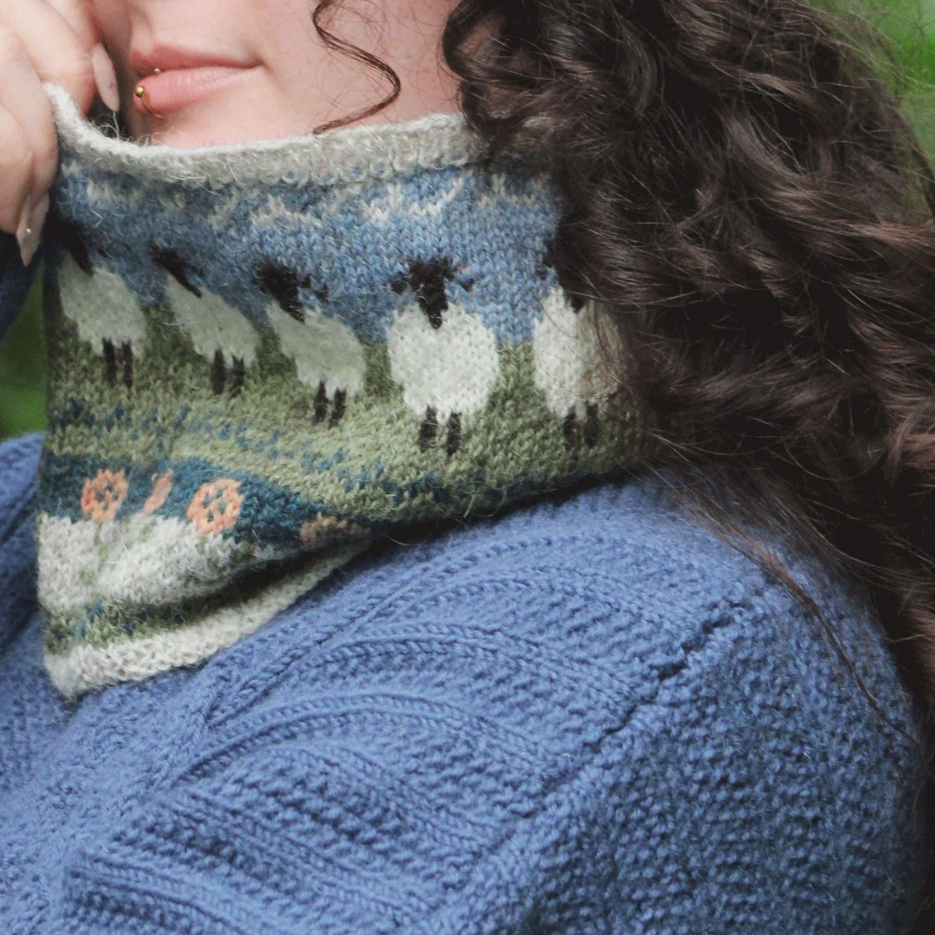 A close up of the knitted cowl, with colourwork of a blue sky, green field and white sheep.