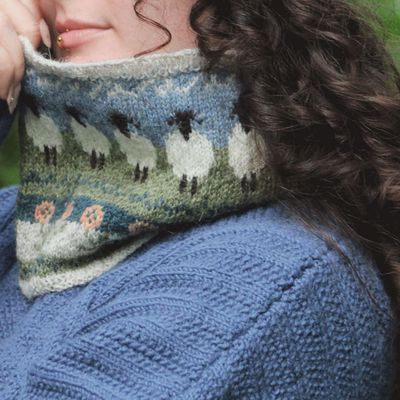 A close up of the knitted cowl, with colourwork of a blue sky, green field and white sheep.