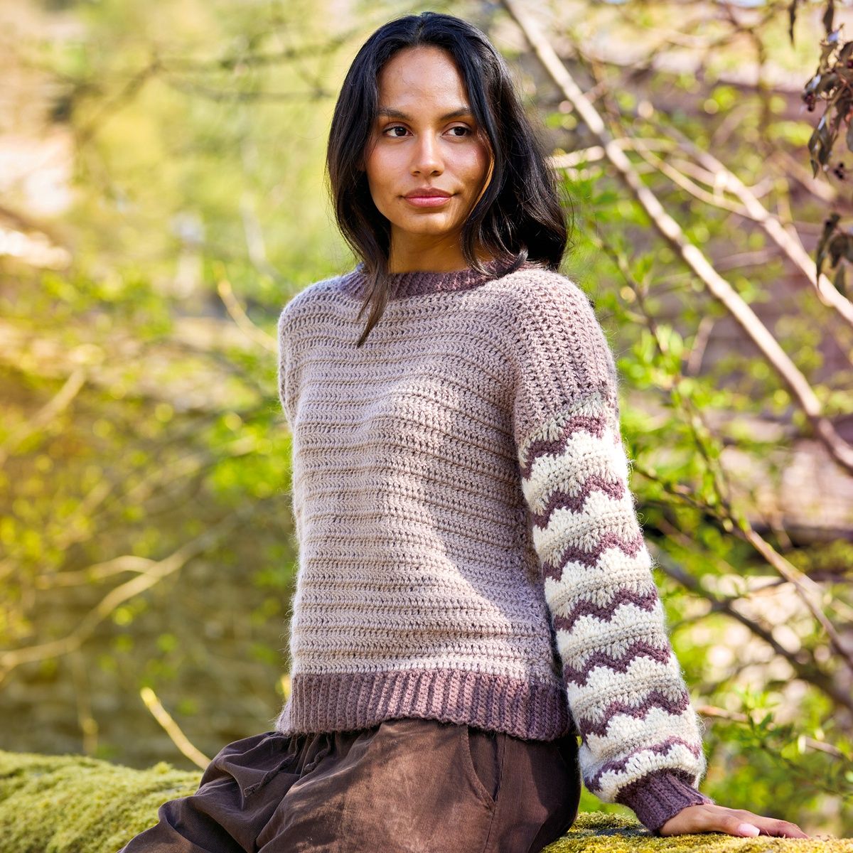 A woman is wearing a pale purple crocheted sweater with purple and cream ripple stitch sleeves.