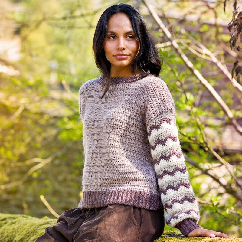 A woman is wearing a pale purple crocheted sweater with purple and cream ripple stitch sleeves.