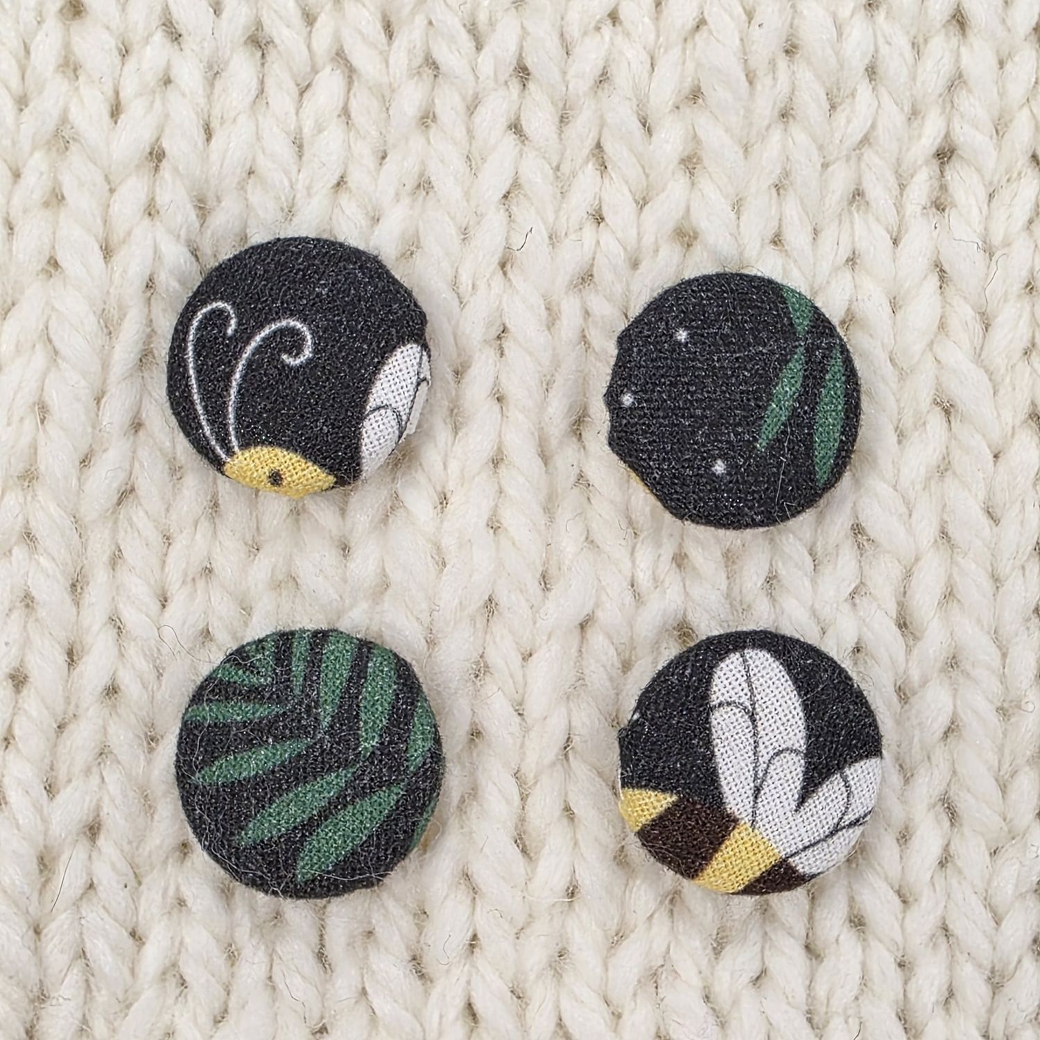 4 black fabric covered buttons with bee and leaf prints, on a cream knitted background.