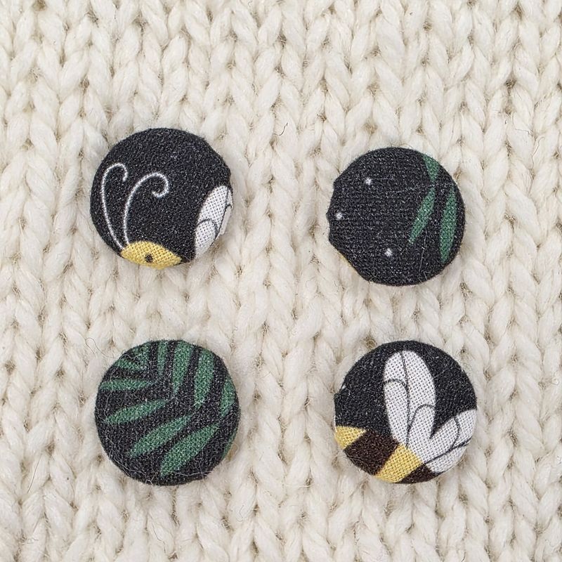 4 black fabric covered buttons with bee and leaf prints, on a cream knitted background.