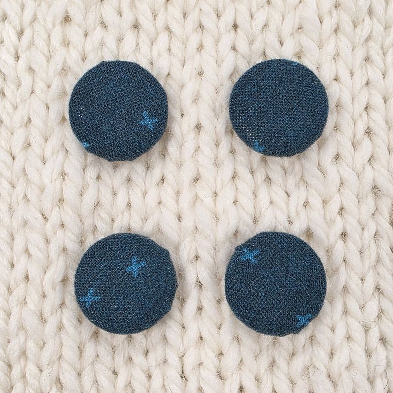 4 navy blue fabric covered buttons with light blue crosses print, on a cream knitted background.