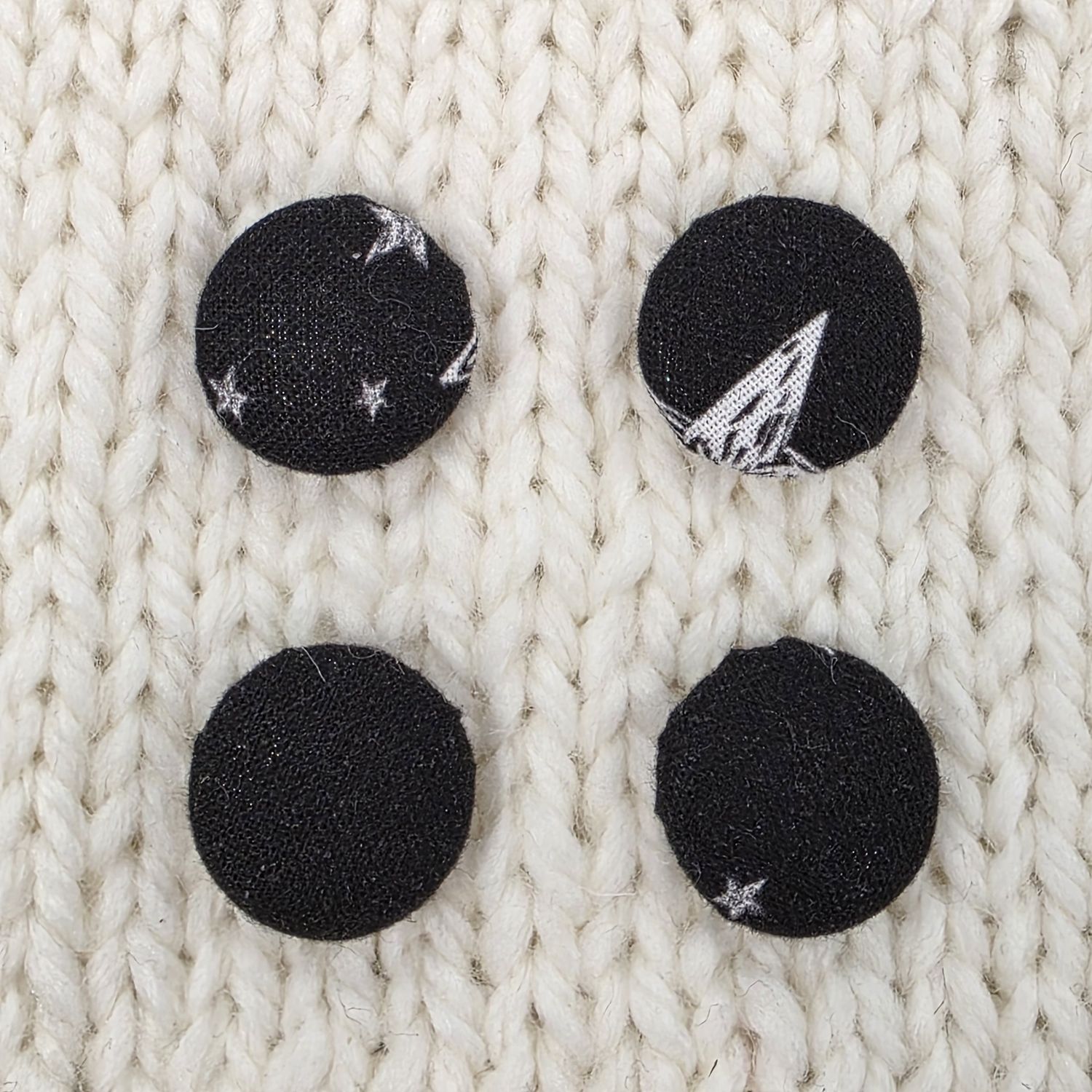 4 black fabric covered buttons with white star print, on a cream knitted background.