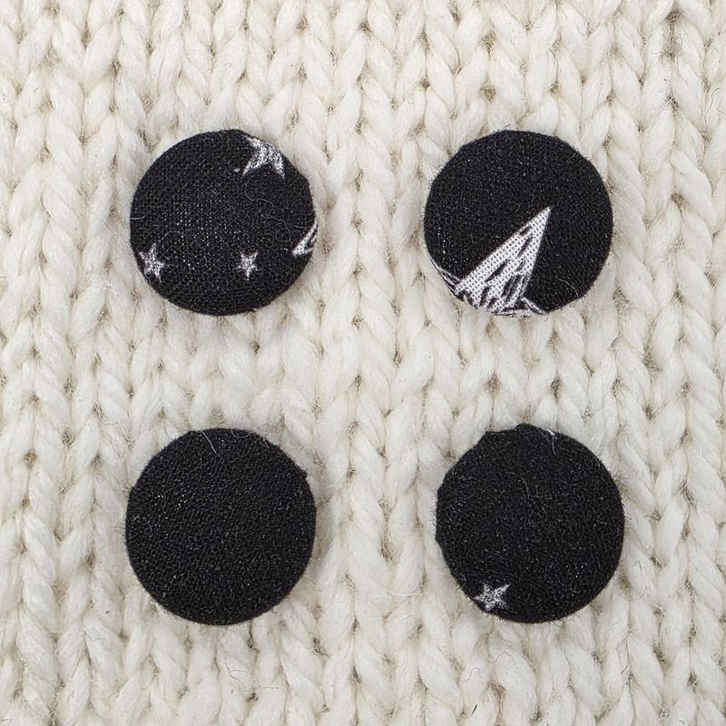 4 black fabric covered buttons with white star print, on a cream knitted background.