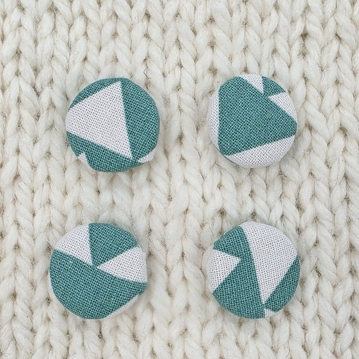 4 white fabric covered buttons with teal green geometric print, on a cream knitted background.