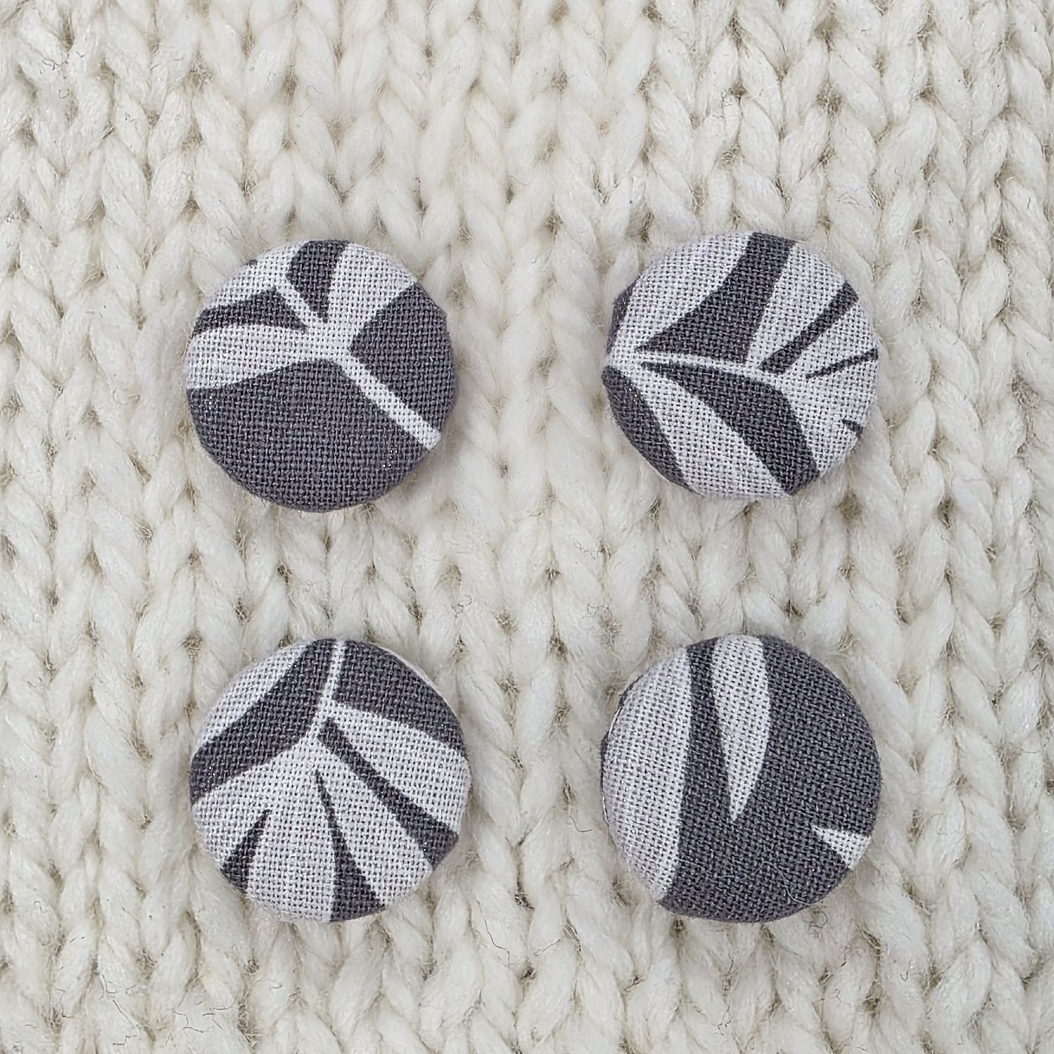 4 dark grey fabric covered buttons with light grey leaf print, on a cream knitted background.