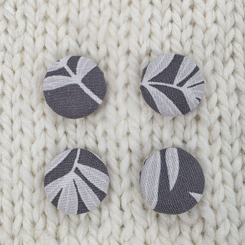 4 dark grey fabric covered buttons with light grey leaf print, on a cream knitted background.