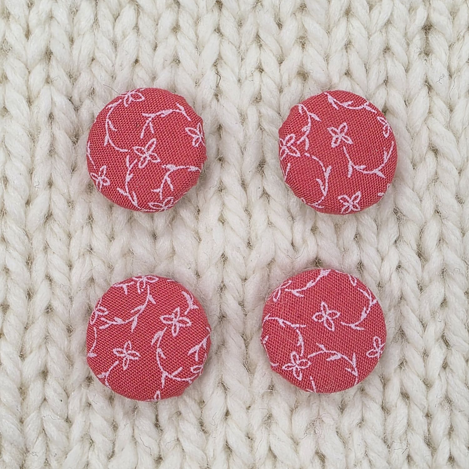 4 coral pink fabric covered buttons with white vine print, on a cream knitted background.