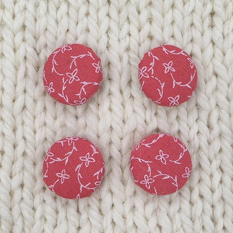 4 coral pink fabric covered buttons with white vine print, on a cream knitted background.
