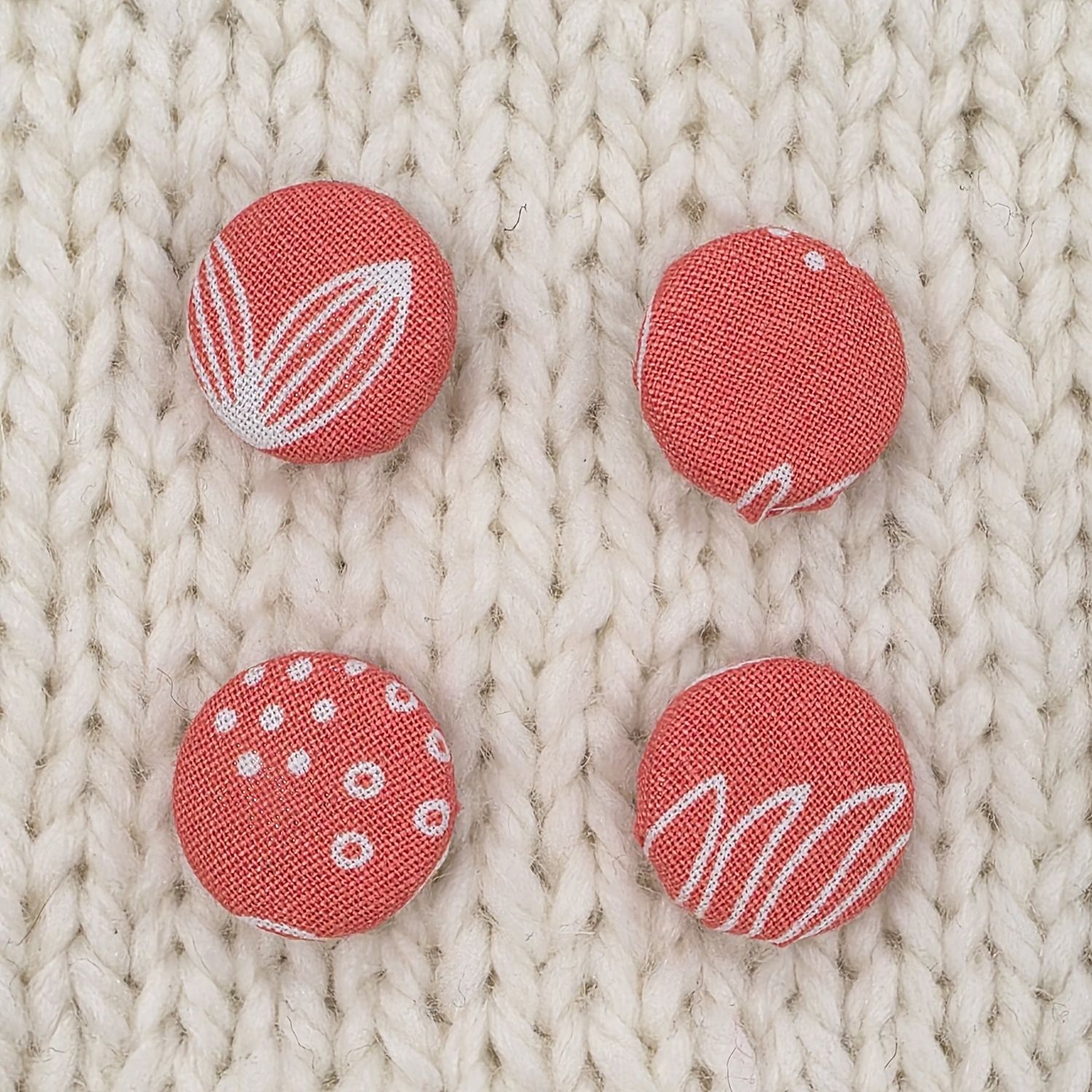 4 coral pink fabric covered buttons with white leaf print, on a cream knitted background.