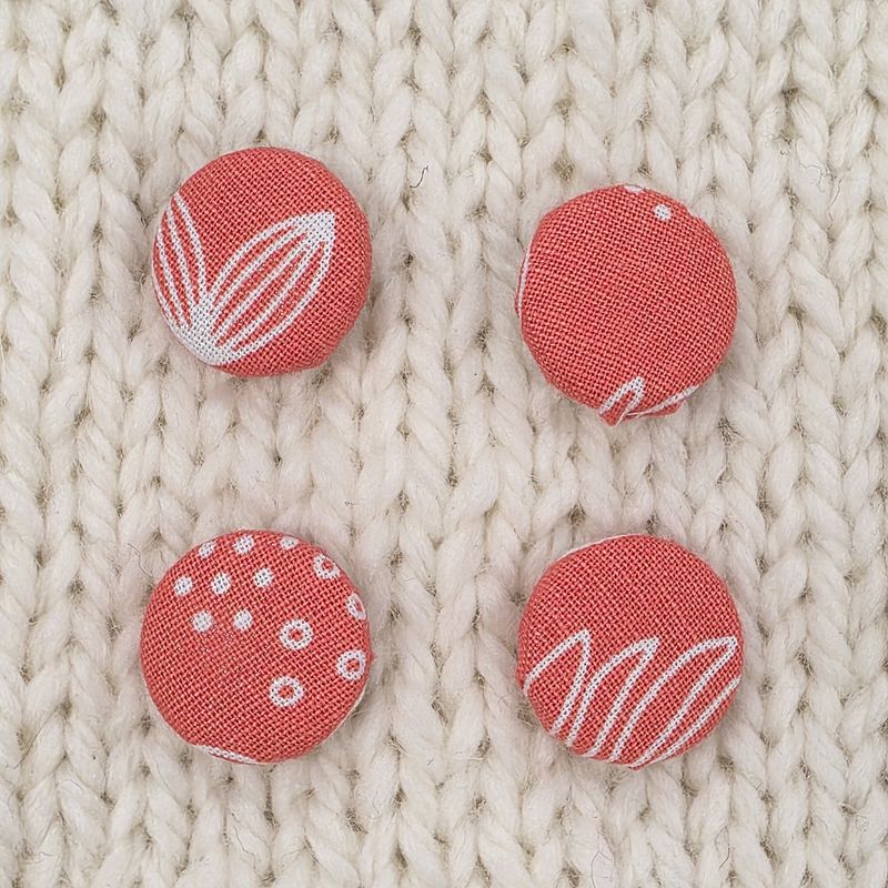 4 coral pink fabric covered buttons with white leaf print, on a cream knitted background.