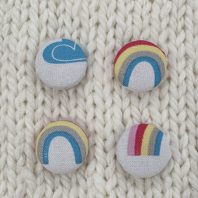 4 white fabric covered buttons with blue cloud and rainbow print, on a cream knitted background.
