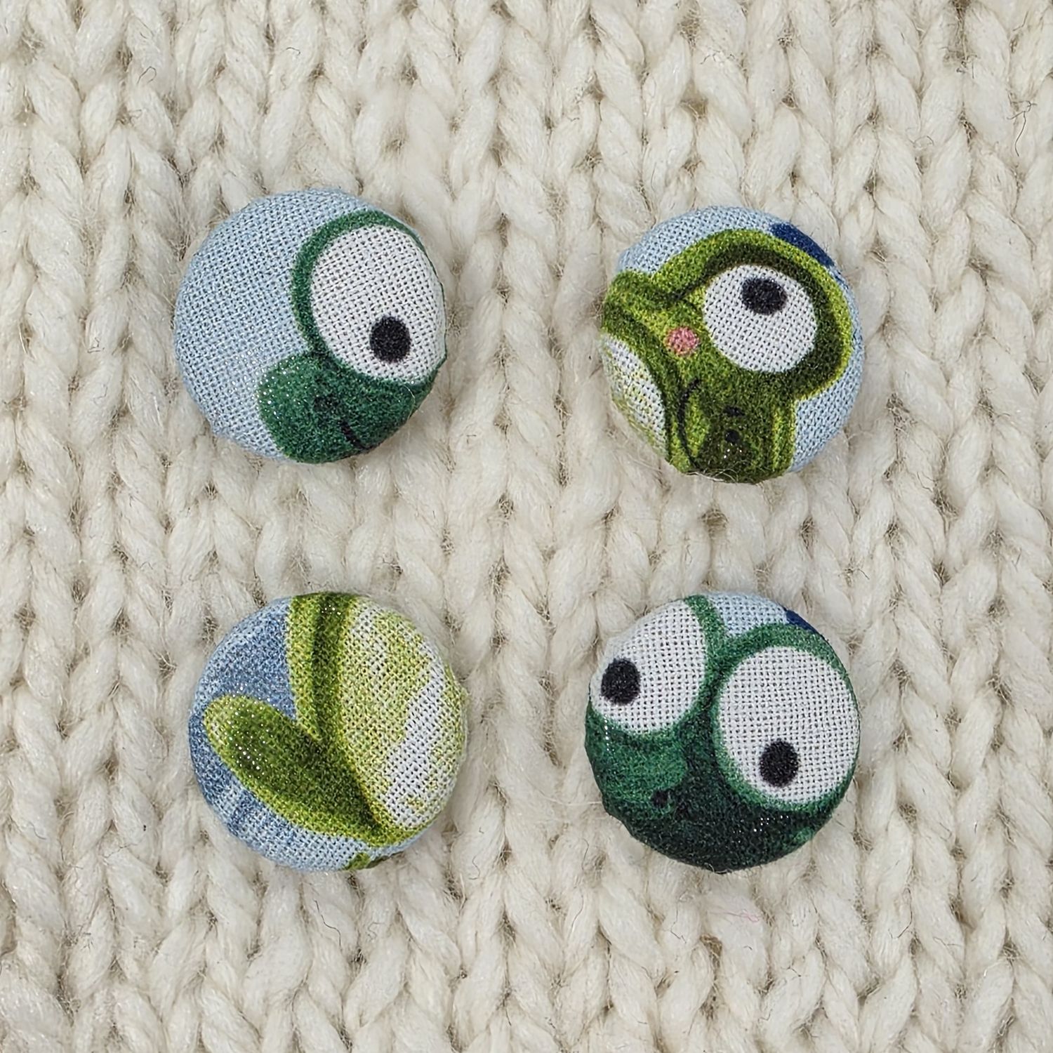 4 blue fabric covered buttons with green frog print, on a cream knitted background.