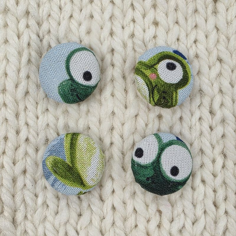 4 blue fabric covered buttons with green frog print, on a cream knitted background.