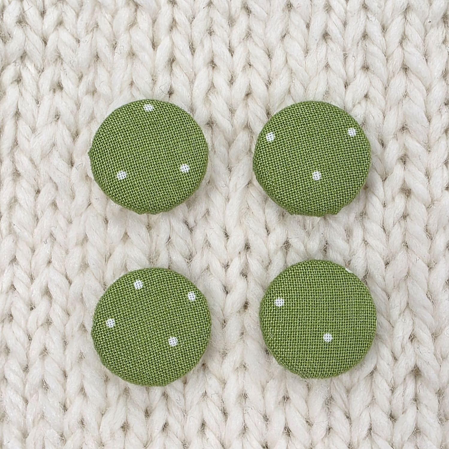 4 green fabric covered buttons with white dotty print, on a cream knitted background.