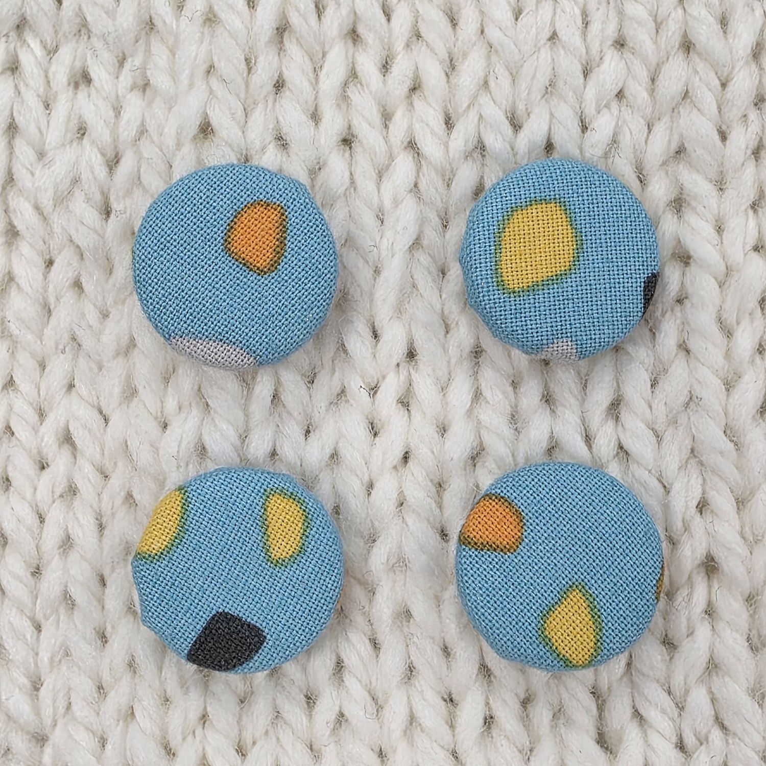 4 blue fabric covered buttons with orange and yellow abstract print, on a cream knitted background.
