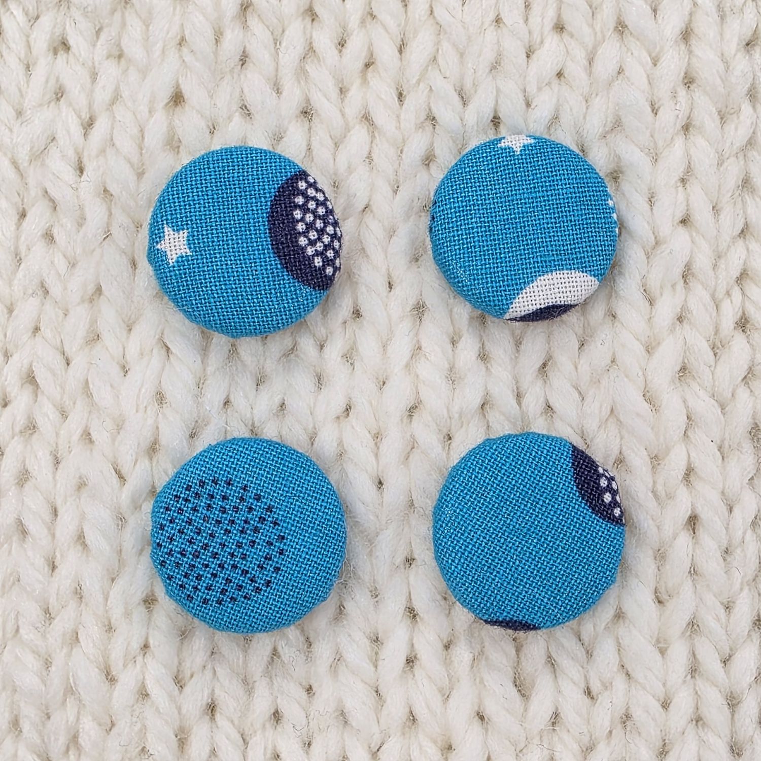 4 bright blue fabric covered buttons with white and navy blue circular print, on a cream knitted background.