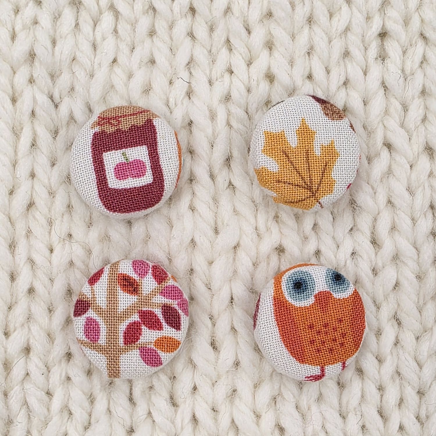 4 white fabric covered buttons with autumnal prints such as a tree, a leaf, an owl and jam, on a cream knitted background.