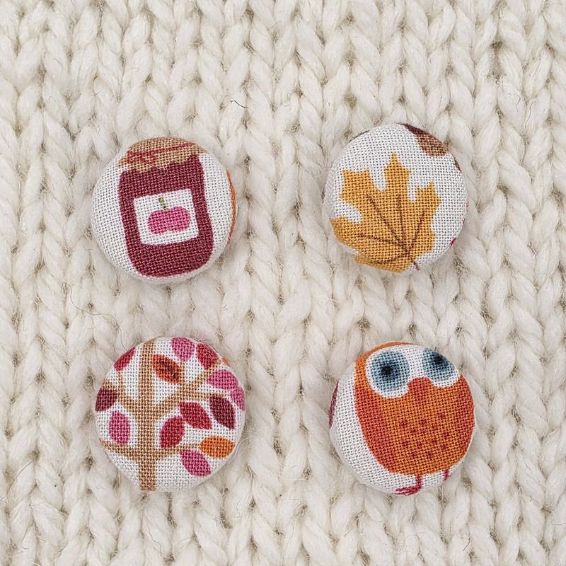 4 white fabric covered buttons with autumnal prints such as a tree, a leaf, an owl and jam, on a cream knitted background.